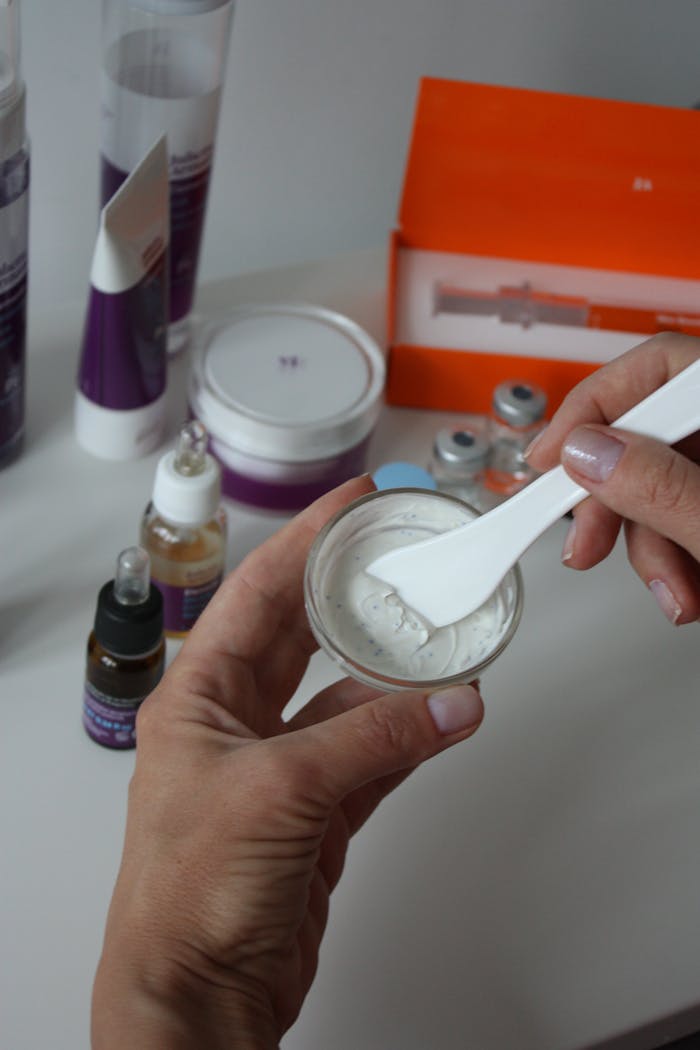 Close-up of hands mixing beauty product in glass jar, surrounded by cosmetics on white background.